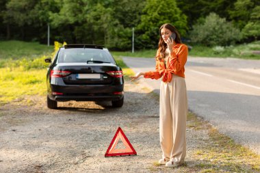 Lady standing near sign of broken car in the middle of the highway and trying to call for help. Car service concept clipart