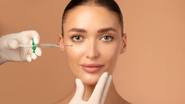 Çekici genç Avrupalı kadın profesyonel klinikte yanağına iğne vuruluyor, kozmetik uzmanı eldivenlerini giyip bayana yüz hatları çiziyor.