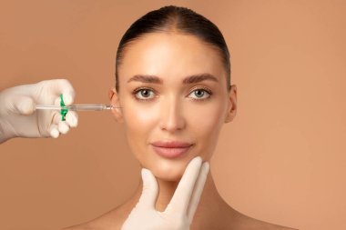 Güzel Avrupalı genç bir kadın güzellik iğnesi yaptırıyor, kozmetik uzmanı elinde şırınga tutuyor, bej arka planı var. Kozmetik ve estetik cerrahi konsepti