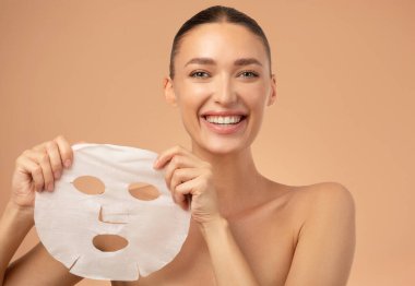 Yüzünde maske olan genç Avrupalı bir kadının bej arka planda kameraya bakıp gülümseyişinin güzel bir portresi. Kozmetik işlem