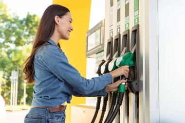 Benzin hortumunu kolunda tutan bir kadın arabasında benzin boşaltmak için dışarıda benzin istasyonunda dikiliyor.