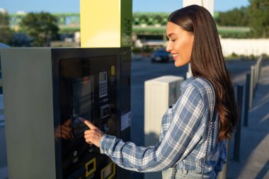 Ekrana dokunan ve park metreyle bilet ödeyen bir kadın. Açık otoparkın yanında duruyor.