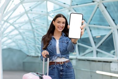 Mutlu bayan havaalanında duruyor ve kameraya boş akıllı telefon ekranı gösteriyor, online bilet rezervasyonu için mobil başvuru öneriyor,