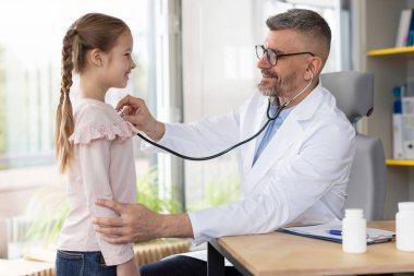 Klinikteki sağlık kontrolü sırasında küçük kızın akciğerlerini kontrol eden erkek doktor, genç bir hastanın nefes alışını incelemek için steteskop kullanan arkadaş canlısı çocuk doktoru.