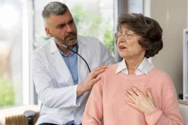 Doktor yaşlı kadın hastayı muayene ediyor, kalp ağrısı çekiyor, erkek doktor hastaya tedavi planını anlatıyor.