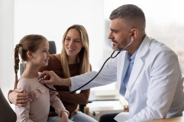 Anne ve kızı çocuk doktorunu ziyaret ediyor, erkek doktor kontrol sırasında steteskoplu küçük bir hastayı muayene ediyor.