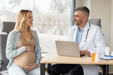 Doktor randevusu. Orta yaşlı bir doktor masadaki tıp ofisinde dizüstü bilgisayarla oturuyor ve hamile bir kadına danışıyor.