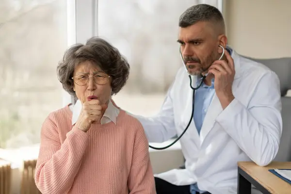 Solunum sistemi kontrol ediliyor. Ciddi bir erkek doktor, öksüren yaşlı kadın hastayı dinliyor, kadın zatürreeden muzdarip, doktor hırıltıyı dinliyor.
