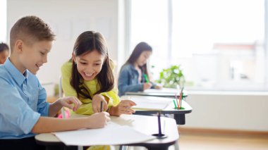 Kız öğrenci, oğlanın yanında oturuyor. Kız, arkadaşının defterinde parmak işareti yapıyor ve gülümsüyor. Sınıfta, panoramada, boş alanda öğrenme sürecinden zevk alıyor.