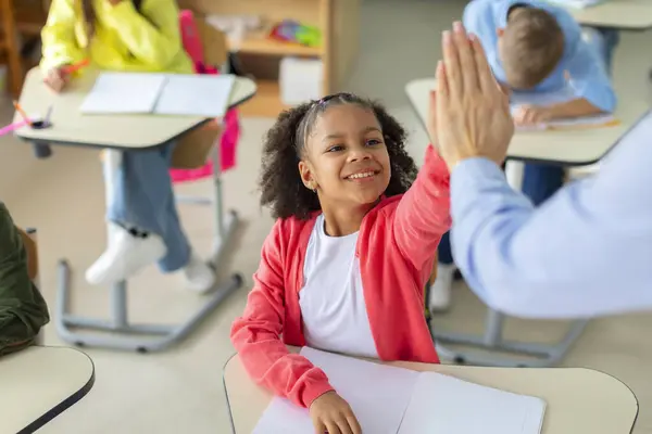 Teacher giving high five to black schoolgirl, achievement, success and mentorship for learning. Students with goals, celebration at school desk
