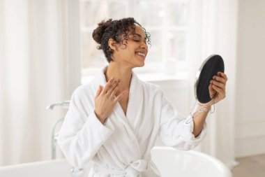 Banyoda mutlu, güzel, siyah bir kadın var ve aynaya bakıyor, boynunu kontrol ediyor, masajdan ve spa tedavisinden sonra sonuçların tadını çıkarıyor.