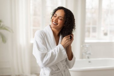 Saç bakım konsepti. Sabah banyodan sonra saçına dokunan bornozlu güzel siyahi kadın banyoda dikilip güzellik rutininin tadını çıkarıyor.