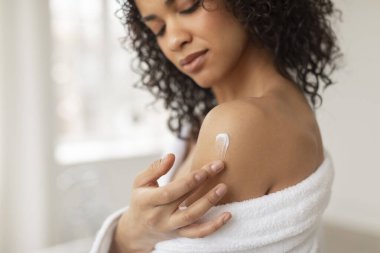 Cilt bakım ürünleri. Genç siyahi kadın duş üstüne nemlendirici krem sürüyor ya da losyon sürüyor banyoda bornoz giyiyor, yakın plan, kesilmiş.