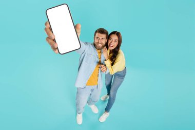 Cep telefonu reklamı. Kameraya boş akıllı telefon gösteren genç çift, turkuaz arka planda tasarım için boş ekran ve kopya alanı gösteriyorlar