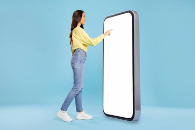 Kadın büyük bir akıllı telefonun önünde duruyor, cep telefonuna dokunuyor boş ekran ve gülümsüyor, en yeni mobil uygulama, mavi arkaplan, kopyalama alanı kullanıyor.