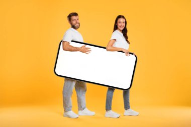 Heyecanlı Avrupalı çift ellerinde büyük bir akıllı telefon ve sarı arka planda beyaz ekranlı bir stüdyo var. Mobil uygulama veya web sitesi tanıtımı için bir model