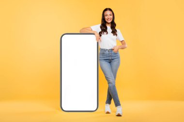 Beyaz ekranlı akıllı telefona yaslanan neşeli genç Avrupalı bayan uygulama, web sitesi veya reklam için modelleme yapıyor, sarı stüdyo geçmişi var.