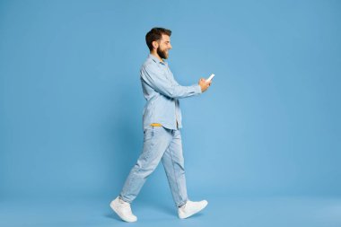Sıradan kıyafetli genç adam akıllı telefonuyla yürüyor ve mavi stüdyo arkaplanı üzerinden mesaj atıyor.