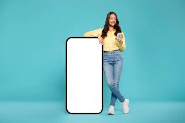 Güzel Avrupalı bir kadın, mavi arka planı beyaz ekranlı, büyük bir cep telefonuyla poz veriyor ve reklam için akıllı telefon kullanıyor.