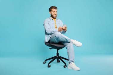 Yakışıklı Avrupalı adam cep telefonu kullanıyor, sandalyede oturuyor, internette ya da sosyal medyada geziniyor, mavi stüdyo arka planındaki yeni mobil uygulamaları kontrol ediyor.