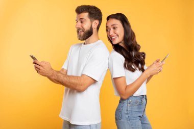 Genç bayan akıllı telefon kullanıyor ve kocasının cep telefonuna bakıyor. Bu arada adam sosyal medyayı karıştırıyor ve gülümsüyor. Sarı stüdyo arka planı.