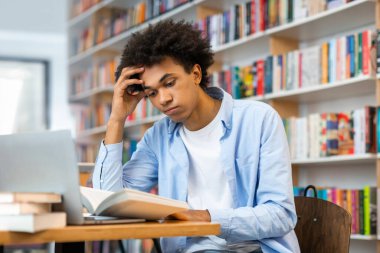 Siyahi erkek öğrenci kampüs kütüphanesinde oturup kitap okuyor, ders vermeye ya da sınava hazırlanıyor. Bilgi ve öğrenme kavramı