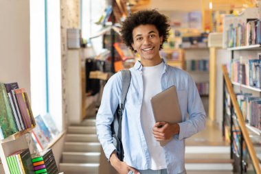 Sırt çantası ve ders kitapları olan Afrikalı Amerikalı bir öğrencinin portresi, ders aralarında üniversite kütüphanesinin yanında duruyor.