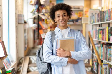 Üniversite kütüphanesinde, etrafı kitaplıklarla çevrili, akademik öğrenim ve araştırma yapan, kameraya gülümseyen gayretli siyah erkek öğrenci.