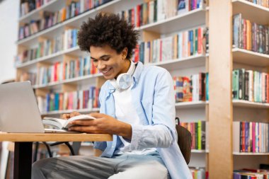 Üniversite kütüphanesinde kitap okuyan siyahi bir öğrenci dizüstü bilgisayarla oturmuş proje hakkında bilgi topluyor.