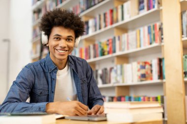 Kolej kütüphanesinin iç kısmında, kulaklık takan ve dizüstü bilgisayar önünde notlar alan mutlu siyahi öğrencinin portresi.