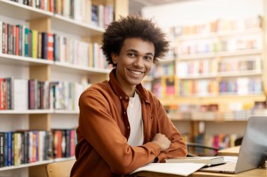 Üniversite kütüphanesinde oturan genç siyahi erkek öğrencinin portresi. Elinde kameraya gülümseyen bir dizüstü bilgisayar reklamı var.