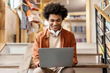 Üniversite kütüphanesinde merdivenlerde oturan, dizüstü bilgisayar kullanan, ders sırasında ara veren siyah bir öğrenci.