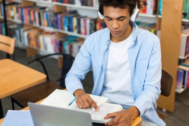 Araştırma ya da okul projesi için kütüphanede dizüstü bilgisayar kullanarak çalışan ve kitap okuyan siyahi bir üniversite öğrencisi.