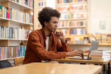 Kitap rafları arasında üniversite kampüsü akademik kütüphanesinde okuyan Afrikalı Amerikalı erkek öğrenci, dizüstü bilgisayar kullanan odaklı adam.