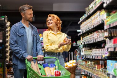 Erkek ve kadın süpermarkette birlikte alışveriş yapıyor, alışveriş yapıyor, yiyecek seçiyor, market koridorunda rafların arasında el arabasıyla yürüyor.