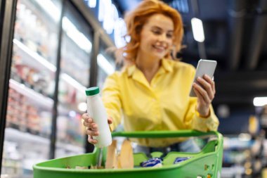 Neşeli kadın müşteri cep telefonundan alışveriş listesini kontrol ediyor ve süpermarketteki arabasına bir şişe süt koyuyor.