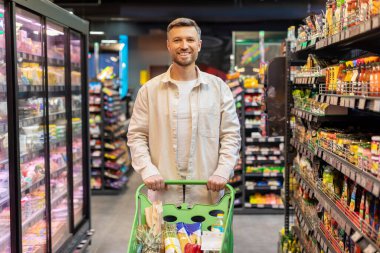Alışveriş arabasını kullanan neşeli Avrupalı adamın portresi süpermarketten alışveriş yaparken kameraya bakıp gülümsüyor..