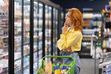 Alışveriş arabalı düşünceli bir bayan markette günlük ürünlerle buzdolabına bakıyor ve ne alacağına karar veriyor. Kadın süpermarketten alışveriş yapıyor..