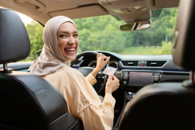 Heyecanlı tesettürlü Müslüman kadın yeni lüks arabayı test ediyor, ön koltukta oturuyor yarı arkasını dönmüş ve kameraya gülümsüyor.