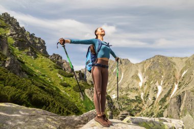 Kaygısız kadın yürüyüşçü ellerini kaldırdı. Dağın tepesinin muhteşem manzarasını kutluyordu. Elinde yürüyüş sopaları ve sırt çantası vardı.
