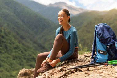 Ayakkabı bağcıklarını bağlayan, dağda kayaların üzerinde oturan mutlu kadın yürüyüşçü, uzağa bakıyor ve gülümsüyor, uzun bir atış. Yazın aktif yaşam tarzı ve aktiviteleri