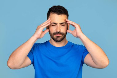 Baş ağrısı olan orta yaşlı bir adam şakaklarına dokunuyor, mavi stüdyo arka planında duygusal bir fotoğraf, boş bir alan. Üzgün bir adam kırmızı alnıyla hafifçe ovuyor, migreni var. Yüksek kalite fotoğraf