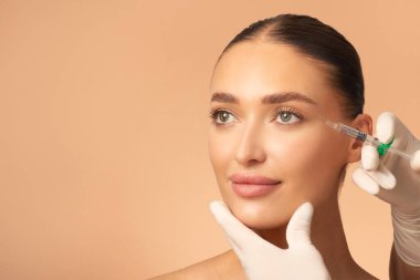 Kozmetik doktoru göz bölgesine kadın iğnesi yapıyor. Kırışıklık önleyici tedavi gören bir kadın. Bej arkaplan ve boş alan.