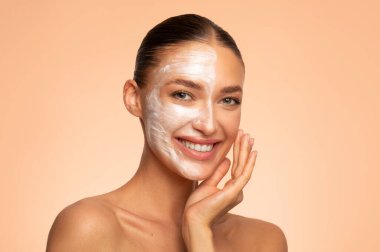 Yüzünün yarısına maske takan, bej arka planda poz veren, kameraya gülümseyen mutlu Avrupalı bir kadının yakın çekim çekimi.