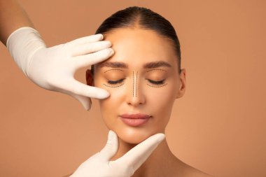 Avrupalı genç bir kadının ön portresi eldivenli estetik cerrahtan konsültasyon alırken, bej renkli stüdyo üzerinde kadın yüzünde izler oluşturuyordu.