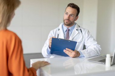 Tıbbi konsültasyon sırasında kadın hastayı dinleyen odaklanmış erkek doktor, klinikte check-up, not panosunda GP yazma reçetesi