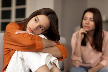 Depresyondaki üzgün genç kız oturmuş dizlerine sarılıyor, annesi bir sonraki kızına bakarken gözlerini kaçırıyor. Ebeveynler ve çocuklar arasındaki iletişim sorunları