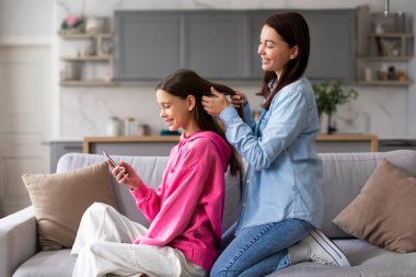 Ergen kızına bakan, saçını tarayan, evdeki kanepede oturan özenli bir anne. Elinde cep telefonu olan genç bir kız annesiyle vakit geçiriyor.