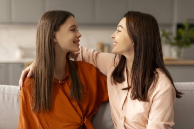 Mutlu anne, genç kızını evinde kucaklıyor, ergen çocuğa sevgisini gösteriyor, birbirinize bakıyor ve kanepede yan yana otururken gülümsüyor.