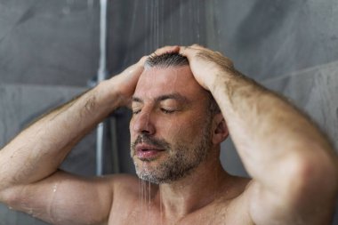 Orta yaşlı bir adam, gözleri suyla kapalıyken banyoda duş alıyor.